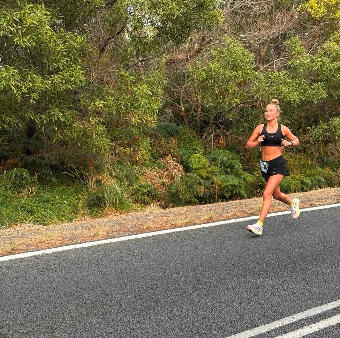 A blonde woman wearing black shorts and a black sports bra running on the road infront of trees 