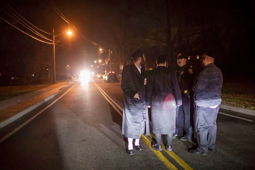 Looking down a straight road lined with double yellow lines at night, you see Orthodox Jewish men talking to a police officer.