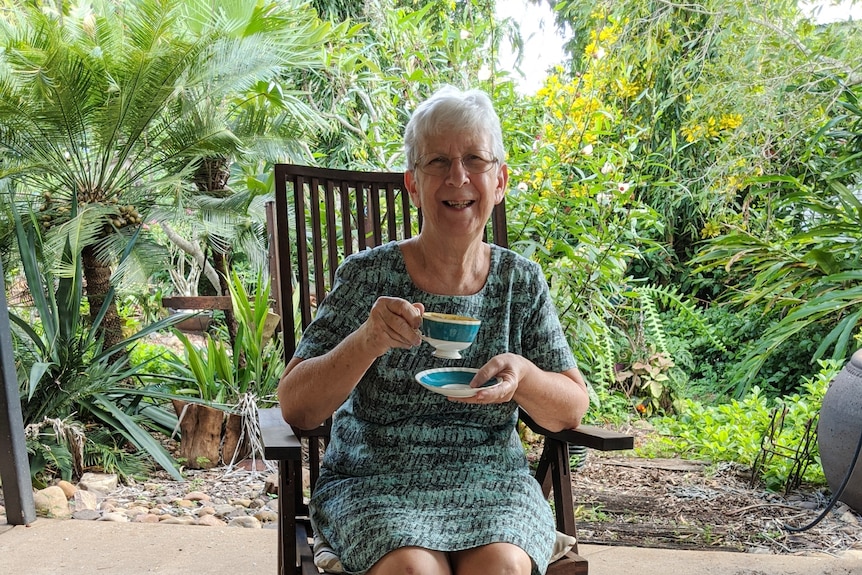 A woman with short grey hair is holding a cup and saucer and smiling. She is sitting outside in a light summer dress.