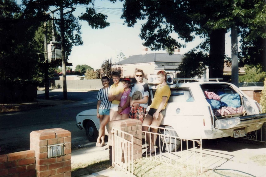 Five people stand against a parked car in a driveway.
