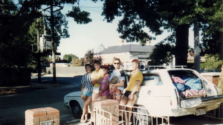 Five people stand against a parked car in a driveway.