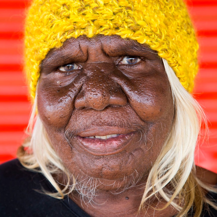 A head shot of Martu elder Nola Taylor wearing a yellow beanie with a red wall in the background.