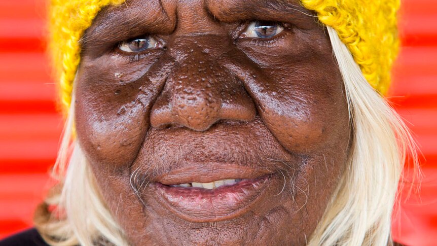 A head shot of Martu elder Nola Taylor wearing a yellow beanie with a red wall in the background.