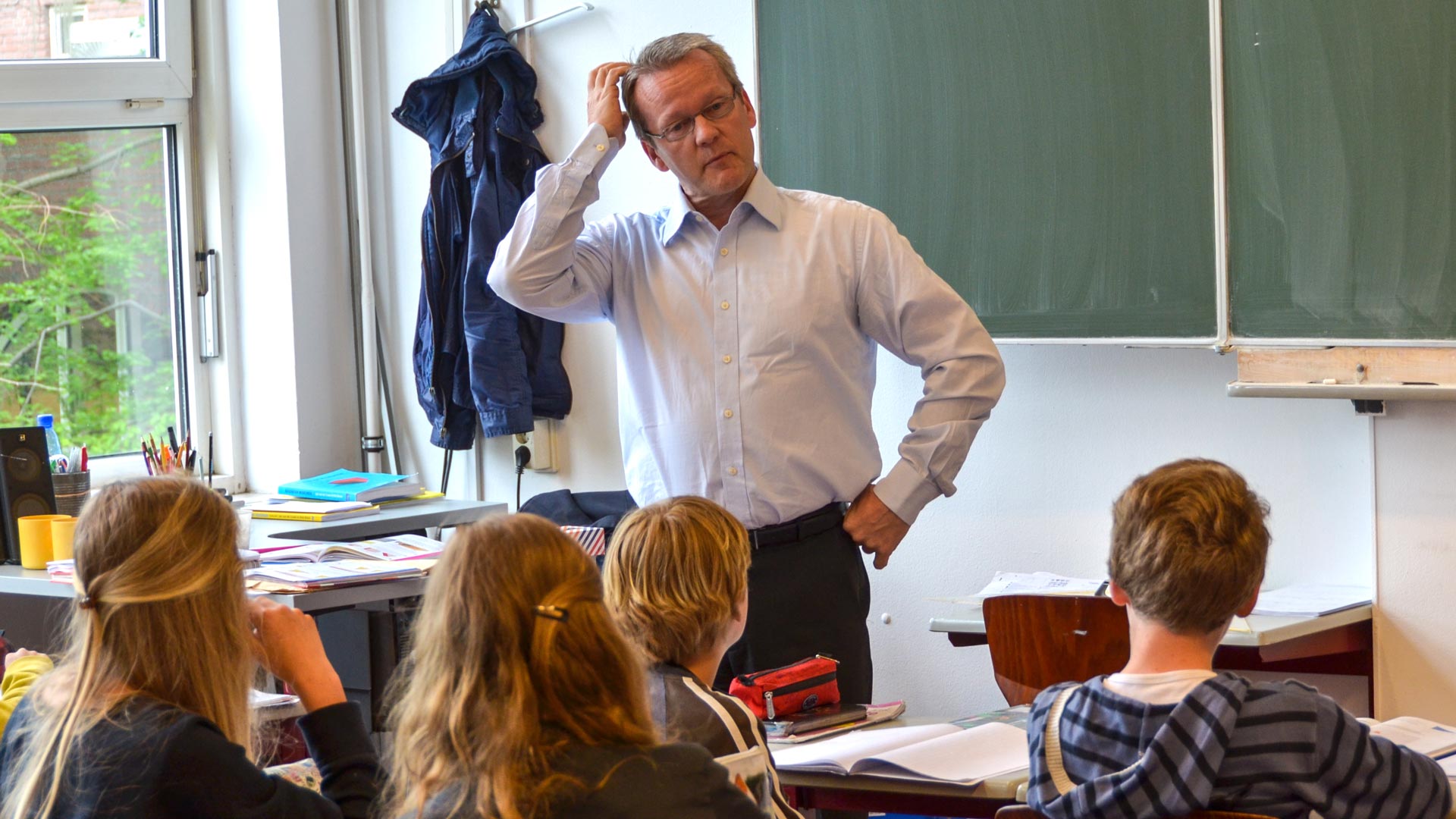 A man stands at the front of a primary classroom and scratches his head as he looks at a young male student.