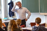 A man stands at the front of a primary classroom and scratches his head as he looks at a young male student.