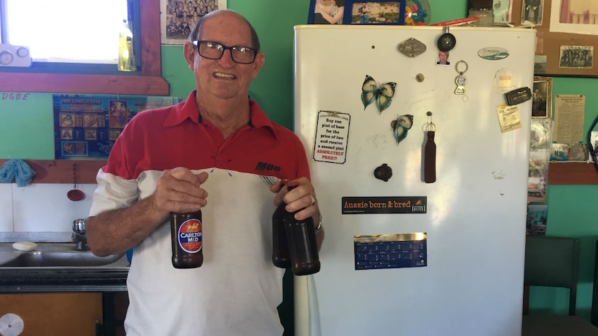A older man with glasses holding three beer bottles, standing in front of a fridge.