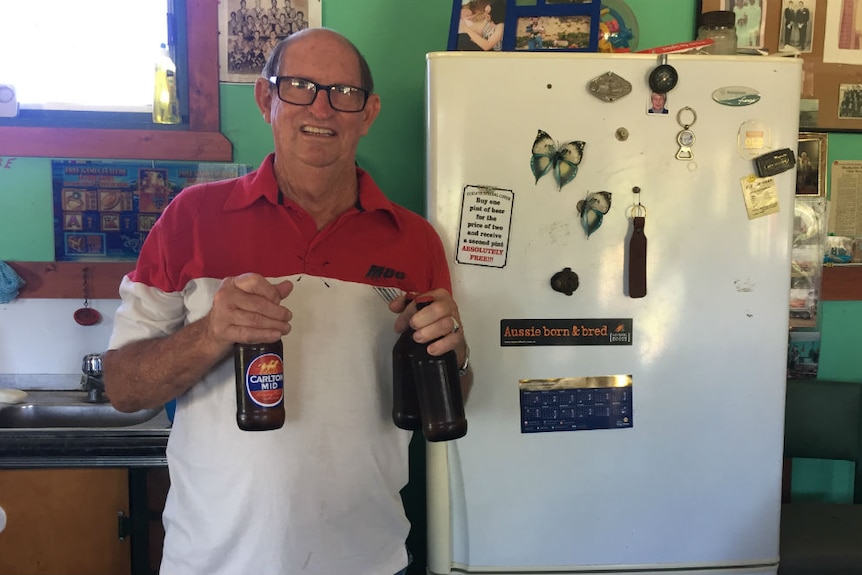 A older man with glasses holding three beer bottles, standing in front of a fridge.