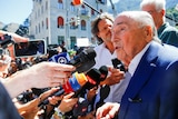 Sepp Blatter wears a suit and talks to a large number of microphones