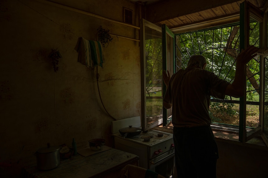 An old man opens a large window. The glass is shattered. The remains of a kitchen in foreground. 