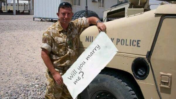 Soldier wearing camouflage gear stands next to tank holding sheet with clearly photoshopped message upon it.