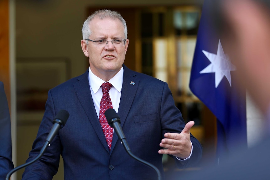 Mr Morrison is looking to the right of frame, and wearing a navy suit and maroon tie. He's standing in front of a flag.