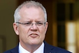 Mr Morrison is looking to the right of frame, and wearing a navy suit and maroon tie. He's standing in front of a flag.