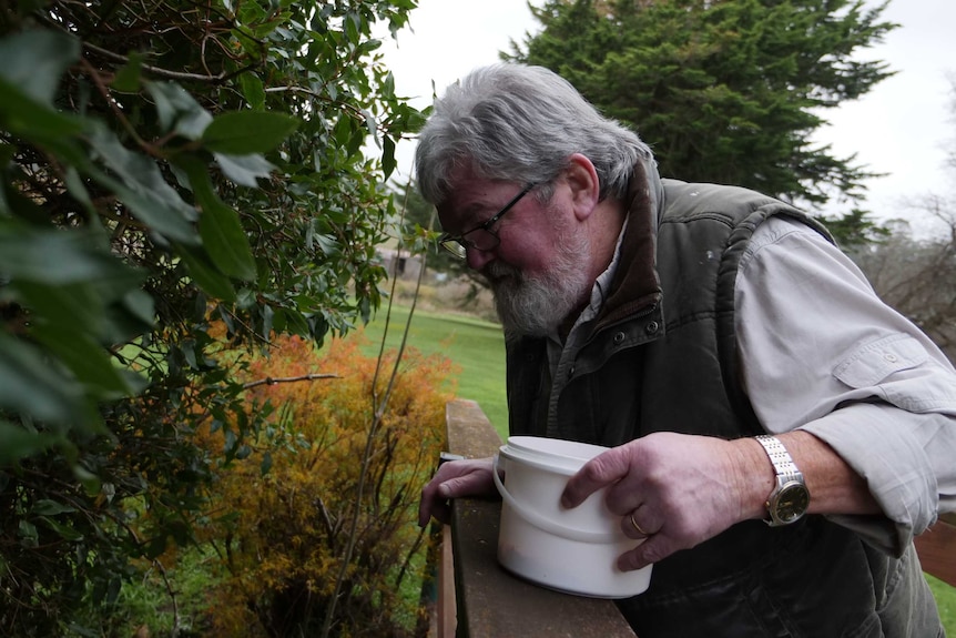 Man with grey hair and glasses with a small bucket in hand, looking down over the side of a small bridge.