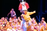 A Samoan woman wearing traditional tapa mat dress and pink woven headdress dances on stage