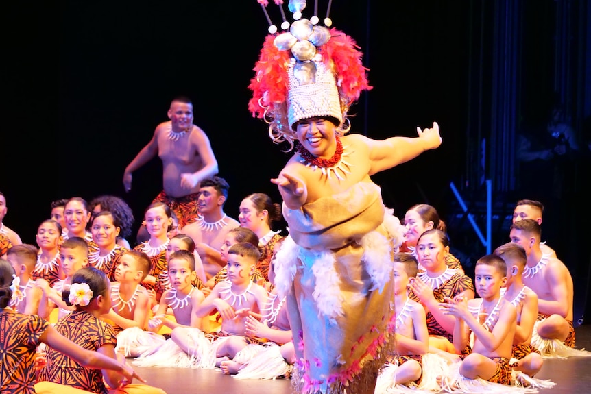 A Samoan woman wearing traditional tapa mat dress and pink woven headdress dances on stage