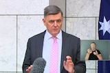 A man in a suit and pink tie standing beside an Australian flag.