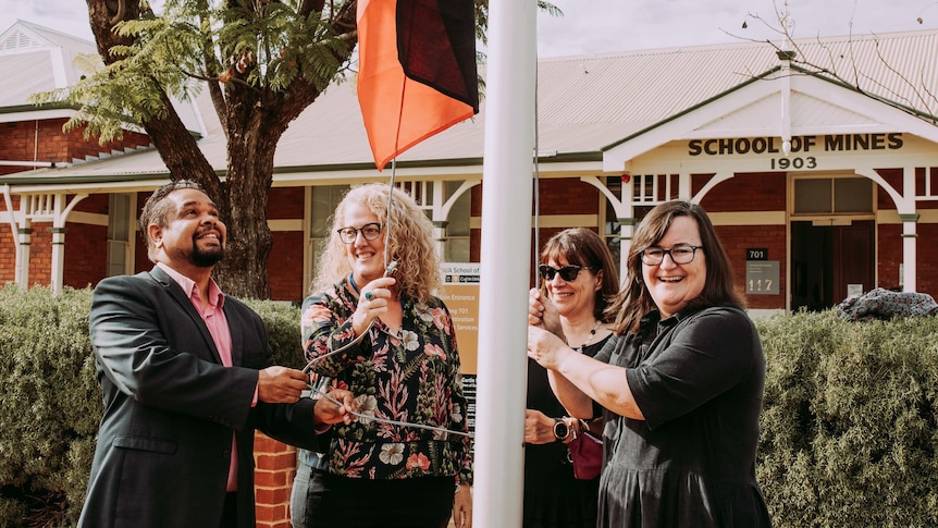 A smiling man and four smiling women raise an Aboriginal flag at a school campus.