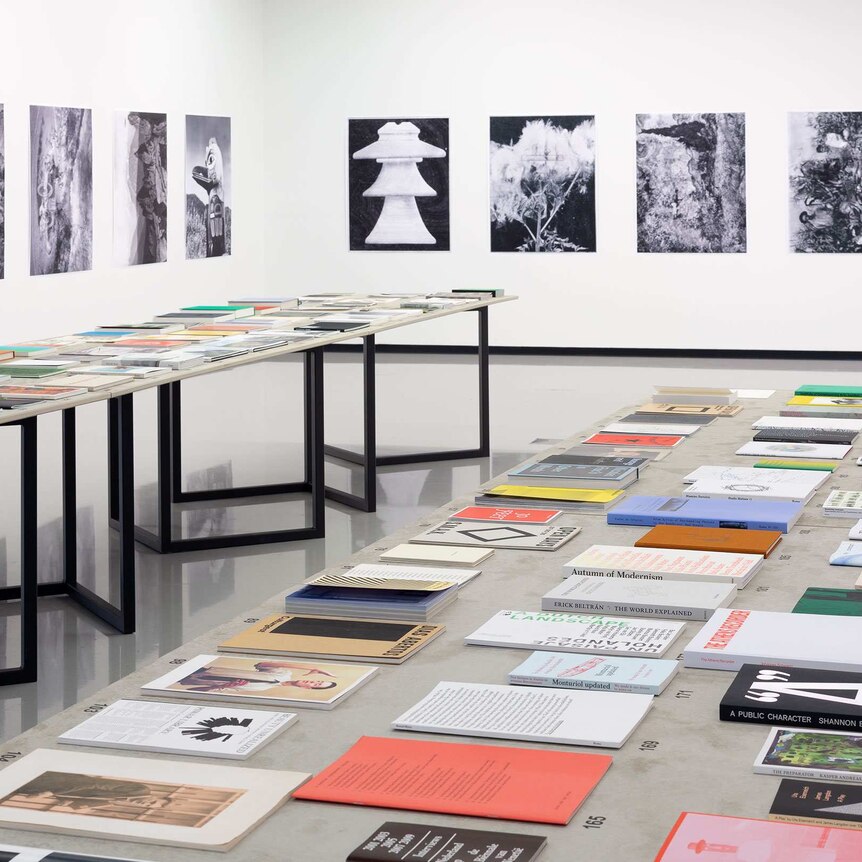 Exhibition view of bookworks: books on the two long tables and black and white prints on the walls