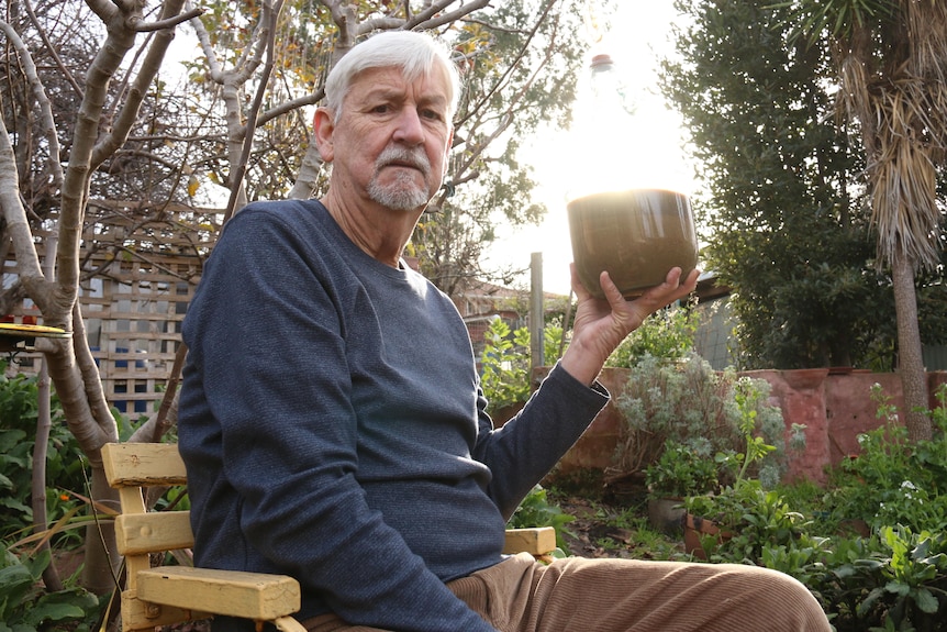 Gerry sits and holds a bottle of brown liquid, outside in his garden.