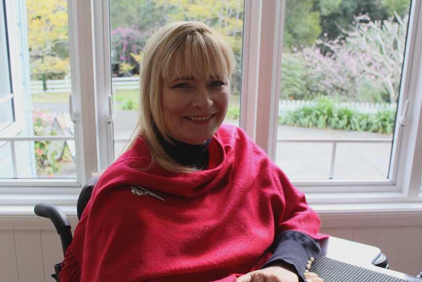 Carol Tayler smiles as she sits in her wheelchair in front of a window wearing a pink scarf