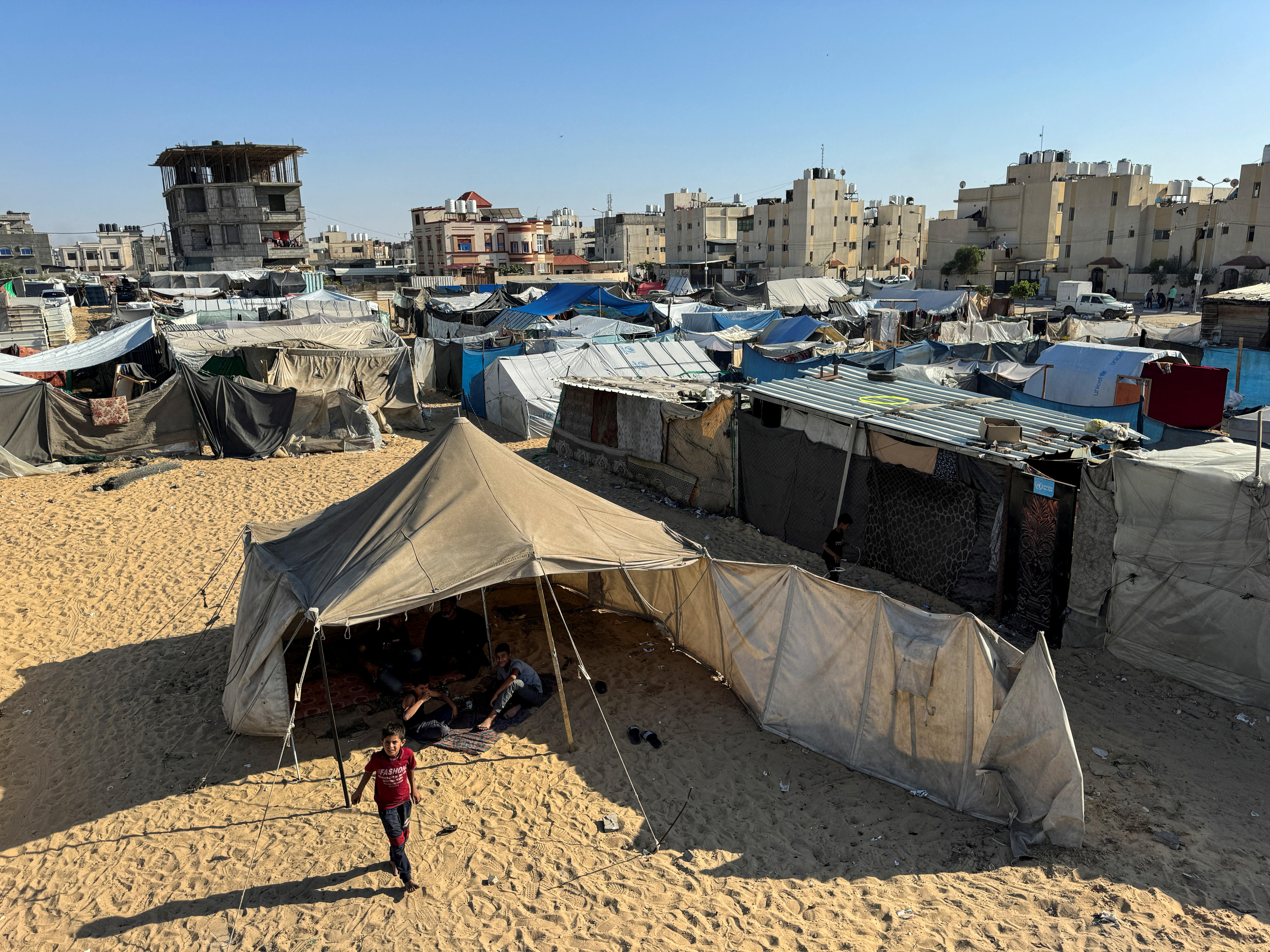 An aerial view of a series of rough-looking tents on a sandy space in front of concrete buildings.