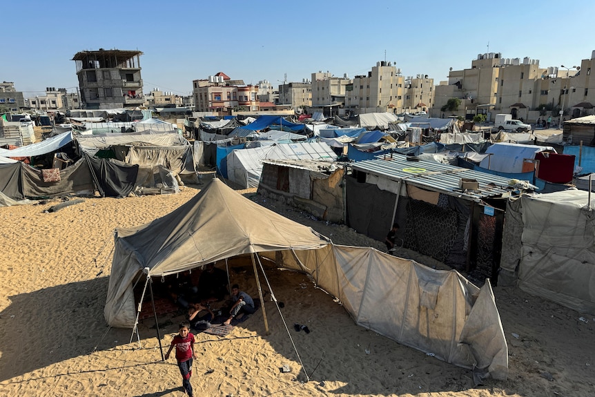 An aerial view of a series of rough-looking tents on a sandy space in front of concrete buildings.