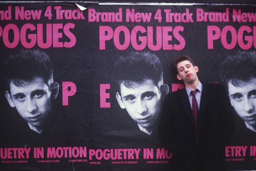 Shane MacGowan wears a suit, stands in front of three The Pogues posters on a street in 1986