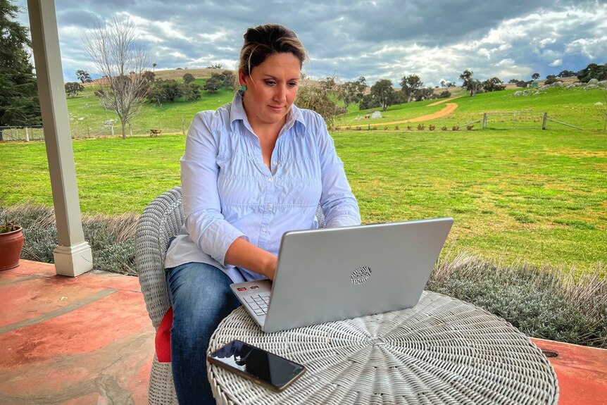 A woman sits on a veranda on a farm typing on a laptop
