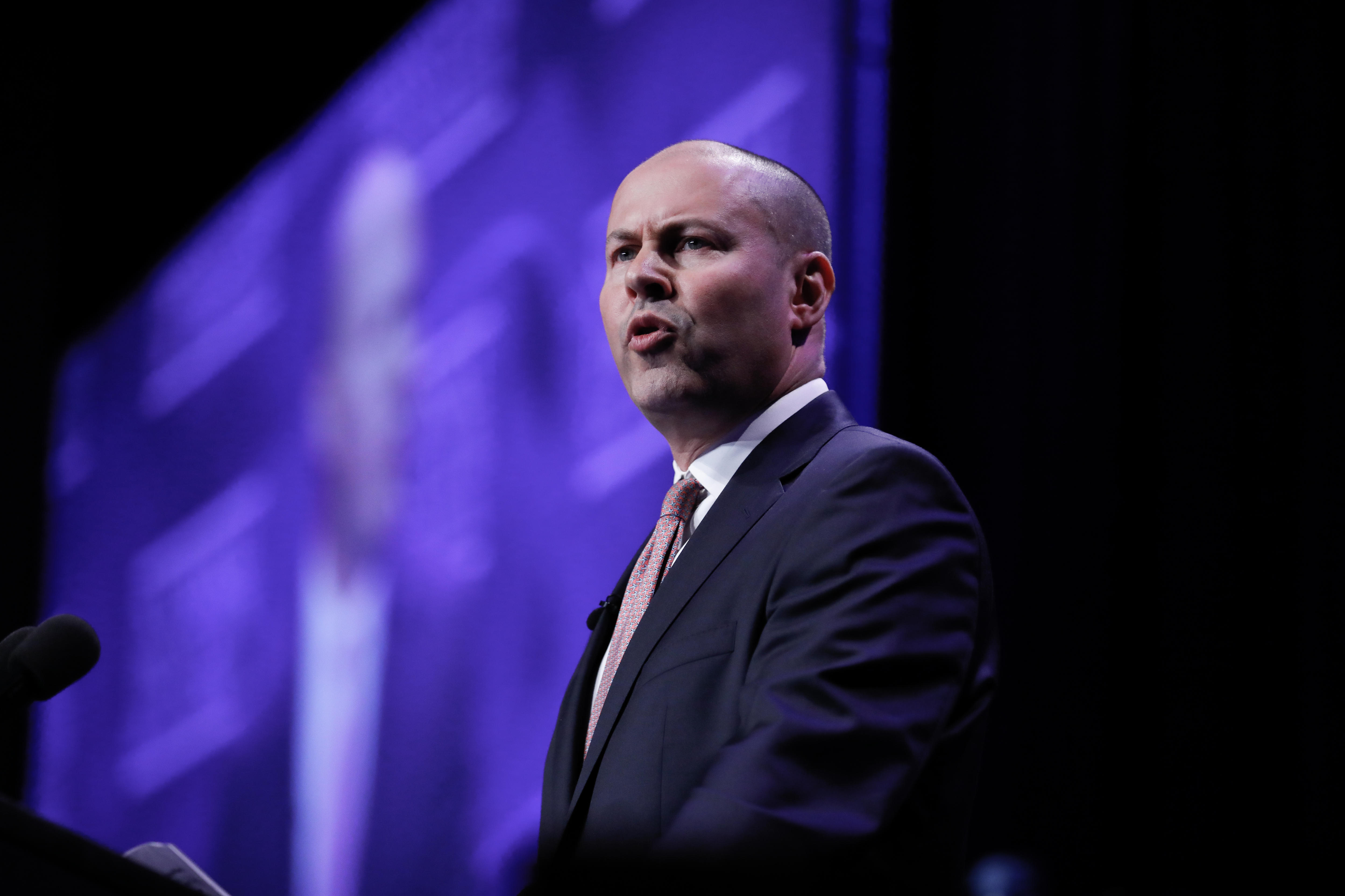 Josh Frydenberg speaks at a lecturn. A screen behind him shows his face