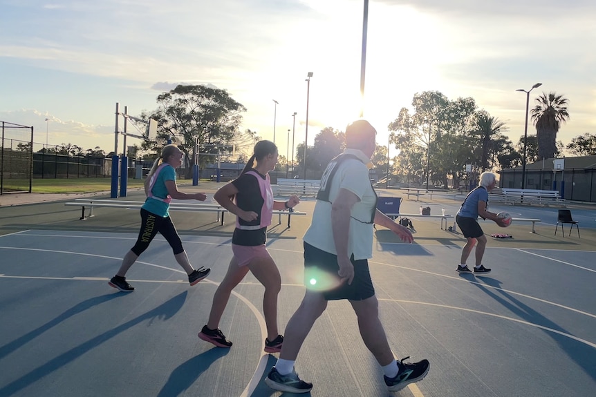 Women aged between 30 and 80 smiling, playing netball outside on a blue court.