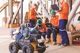 Workers on a mine site control a small, wheeled robot using a laptop