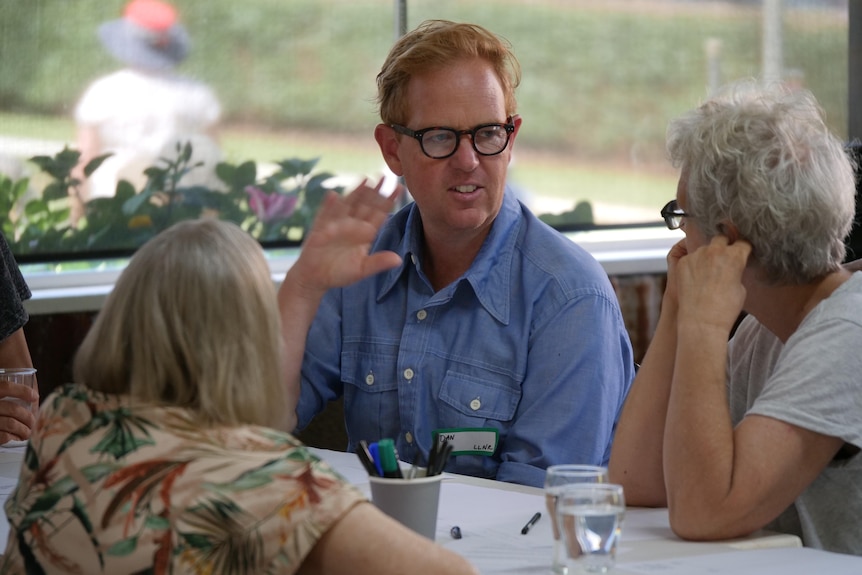 A man seated at a table with other people gesticulates as he talks.