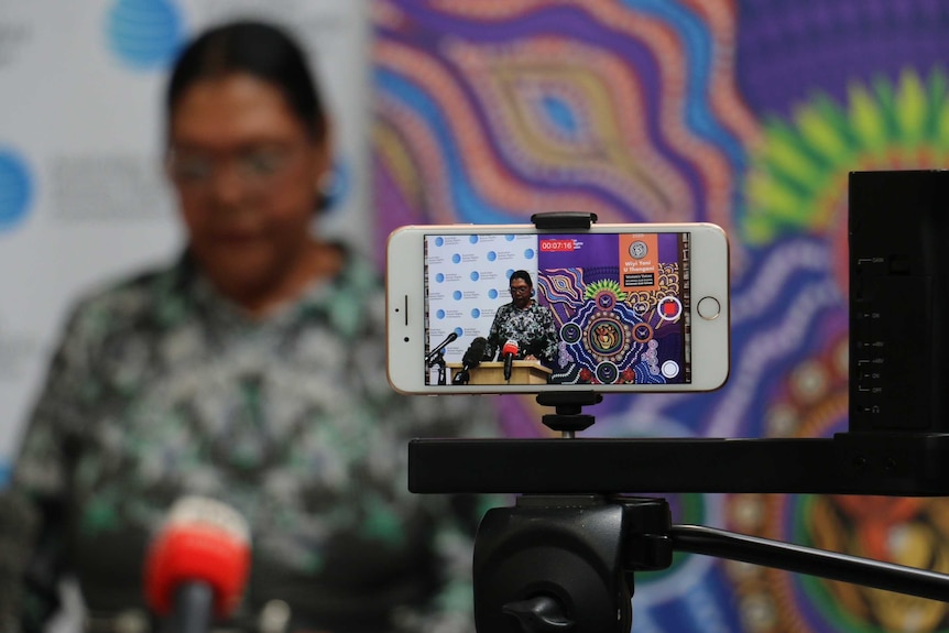 A mobile phone recording June's speech is in focus, with June blurred in the background.