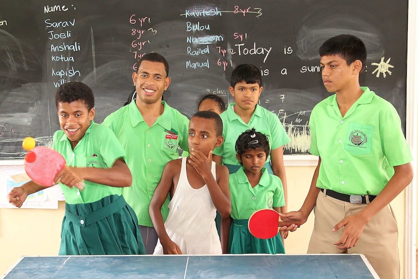 A girl laughs as she hits a ping pong ball as many classmates gather around her.