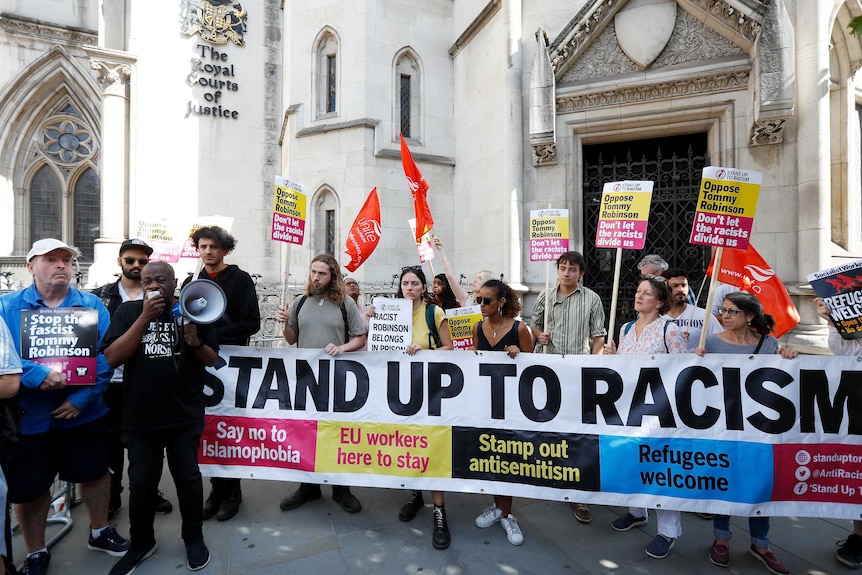 Protesters hold up placards saying "Oppose Tommy Robinson" and hold a banner saying "Stand up to racism".