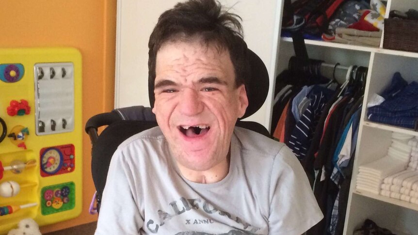 Severely disabled man Colin Burchell smiles at the camera while sitting on a mobility chair in his orange bedroom