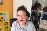 Severely disabled man Colin Burchell smiles at the camera while sitting on a mobility chair in his orange bedroom