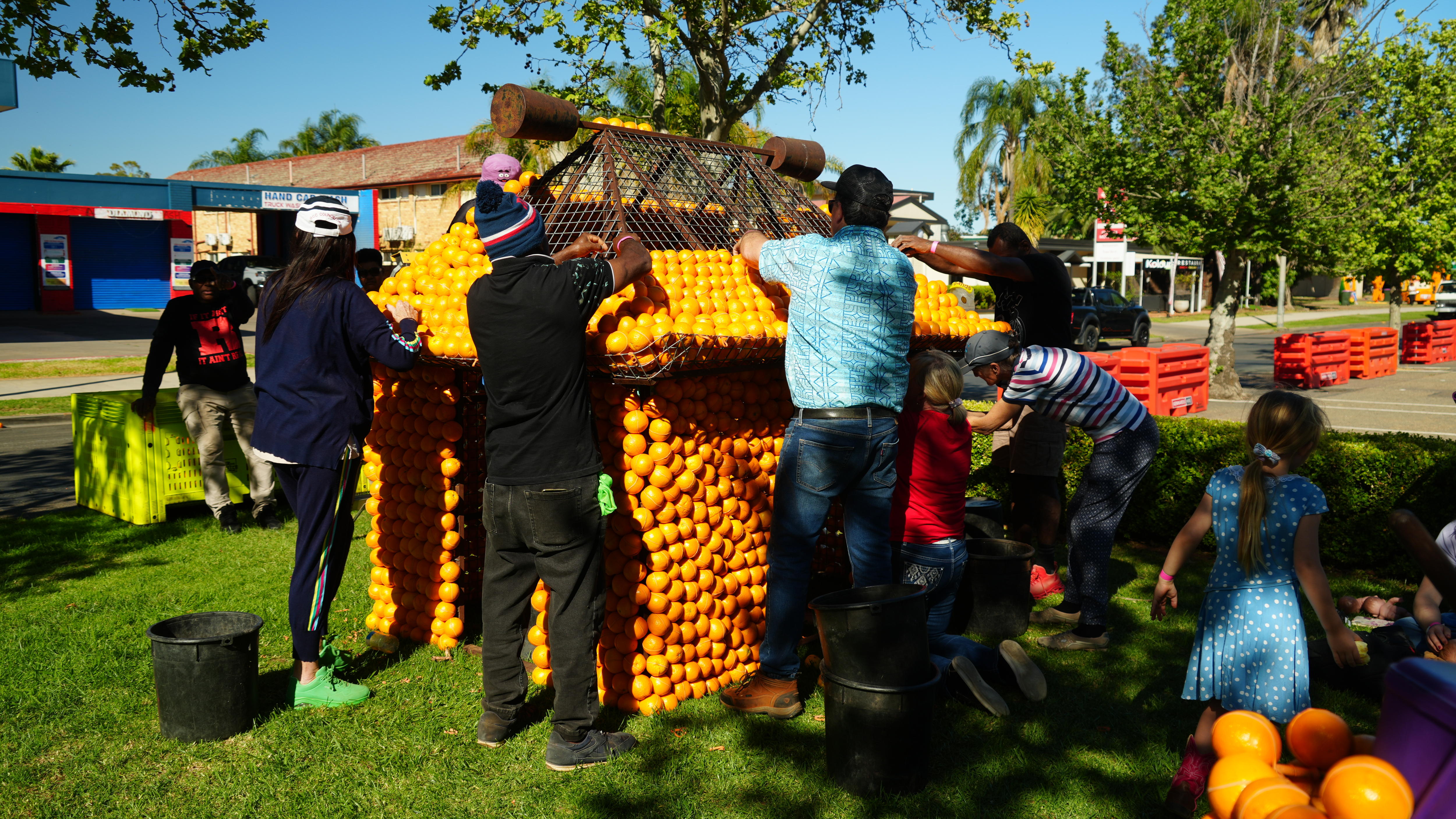 Men attach oranges to a replica of a house on some grass beneath a clear sky.