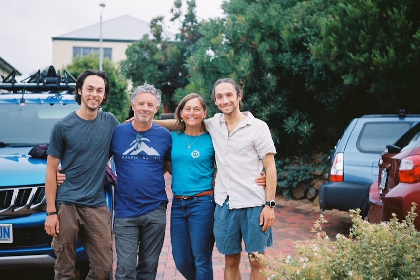 A family — two adult sons and a husband and wife — wrap their arms around each other in a driveway. 