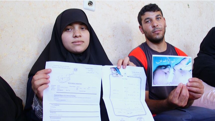 A grieving couple sit leaning against a wall holding a photo of their sick newborn baby and a referral report for his treatment.