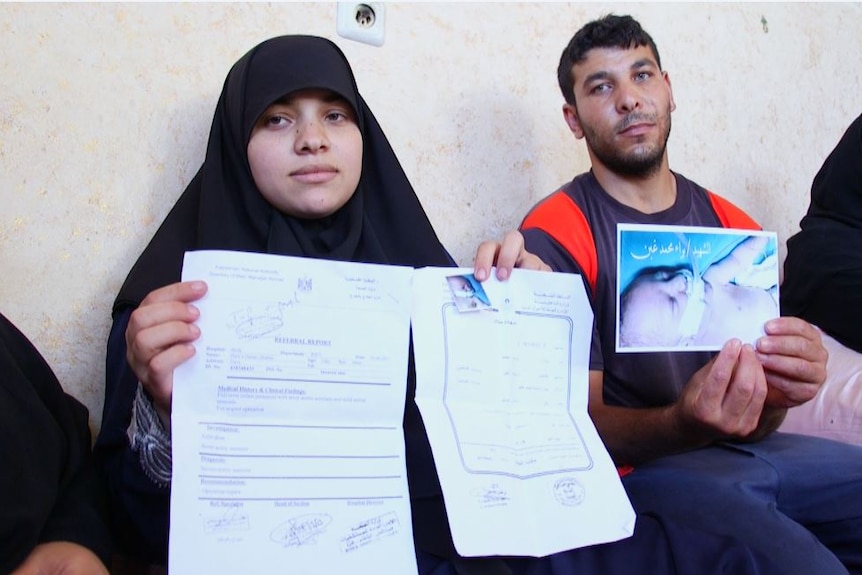 A grieving couple sit leaning against a wall holding a photo of their sick newborn baby and a referral report for his treatment.