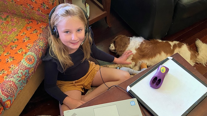 A girl sits on the floor, patting a dog, wearing a head set in front of a computer and paper 