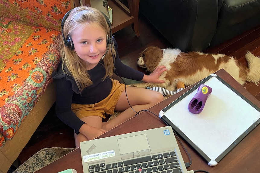 A girl sits on the floor, patting a dog, wearing a head set in front of a computer and paper 