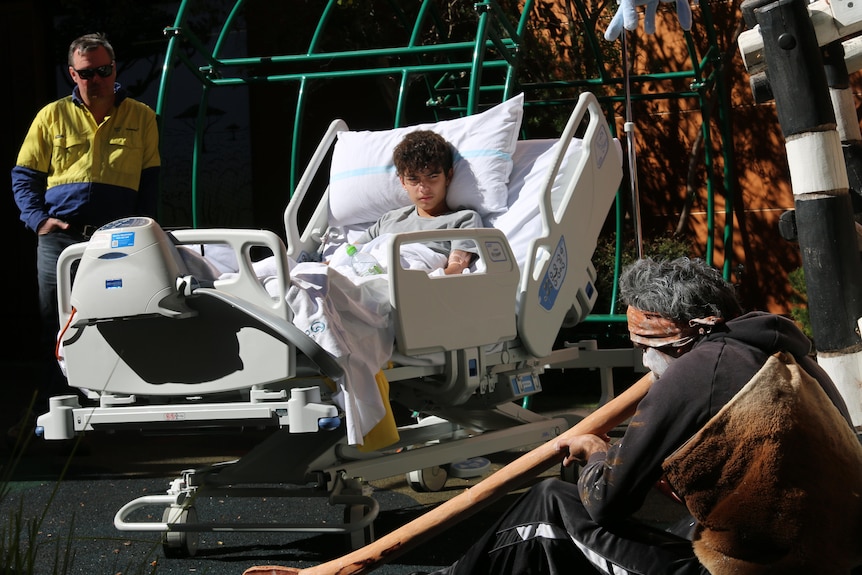 A boy in a hospital bed watches an aboriginal man playing a didgeridoo outside.
