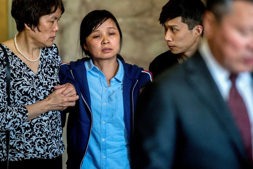 A woman stands supported by two men with her face downcast.