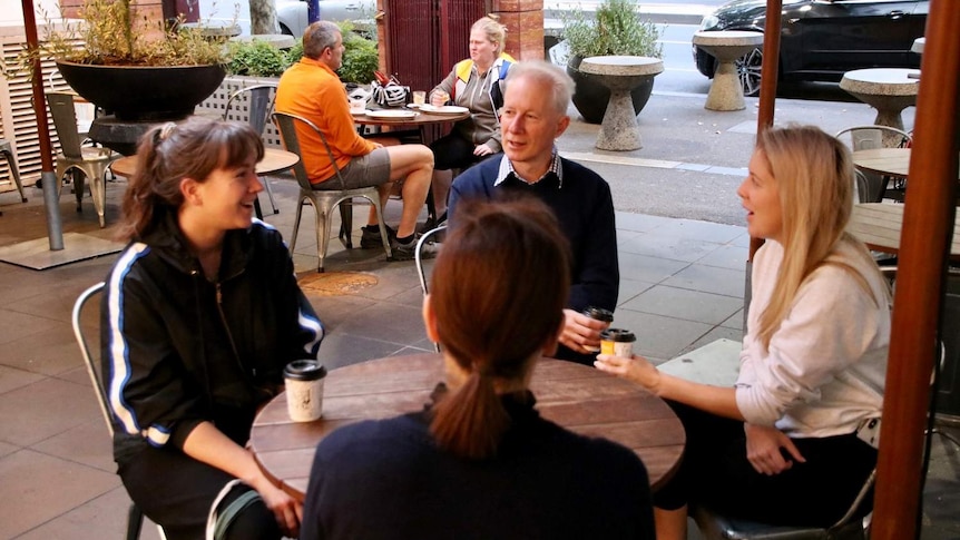 A group of four sit around a table outside at a cafe with takeaway coffee on a bright morning.