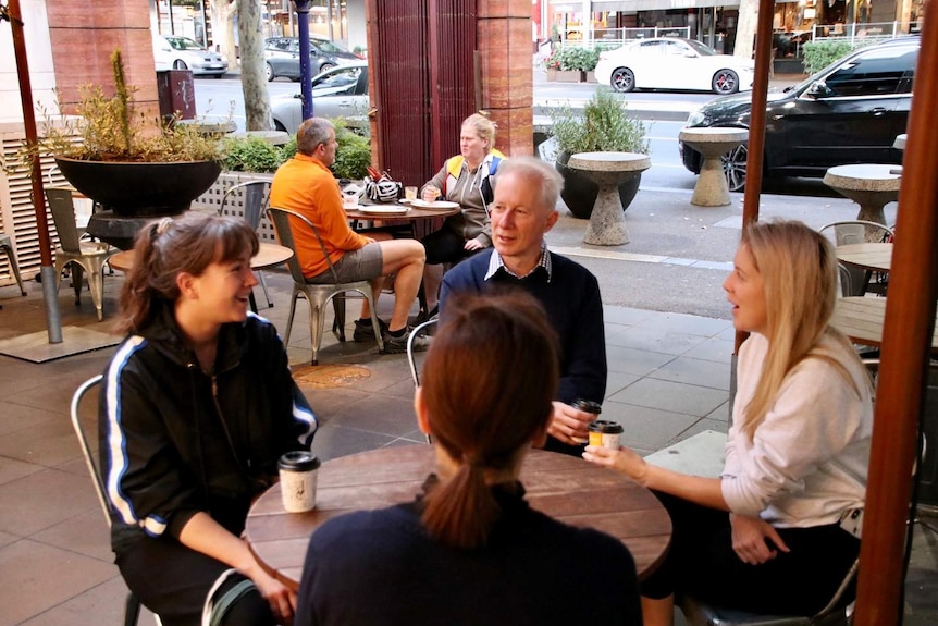 A group of four sit around a table outside at a cafe with takeaway coffee on a bright morning.