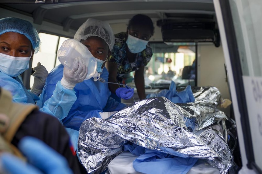 Two women in hospital gowns administer fluids to a burns victim as they lay under foil in the back of a van