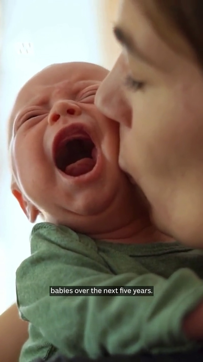 a tight shot shows a woman kissing a baby dressed in green with its mouth open