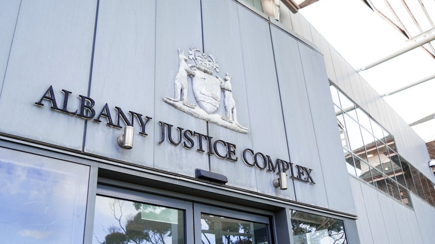The exterior of a building, with glass doors and stone visible, and a sign that reads Albany Justice Complex 